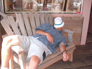 Ein Mann schläft auf einer Holzbank mit einem Hut über dem Gesicht. Offensichtlich Frühjahrsmüdigkeit, denn es ist Tag hell.