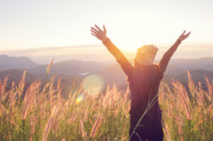 Eine Person blickt dem Sonnenuntergang entgegen und streckt die Arme vital nach oben.